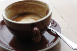Brown cup of coffee on a matching saucer, with a teaspoon resting beside it on a light wooden table.